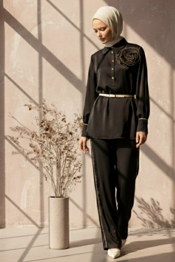 Woman in a  modest black satin pant set with a hijab standing in a sunlit room with a plant.