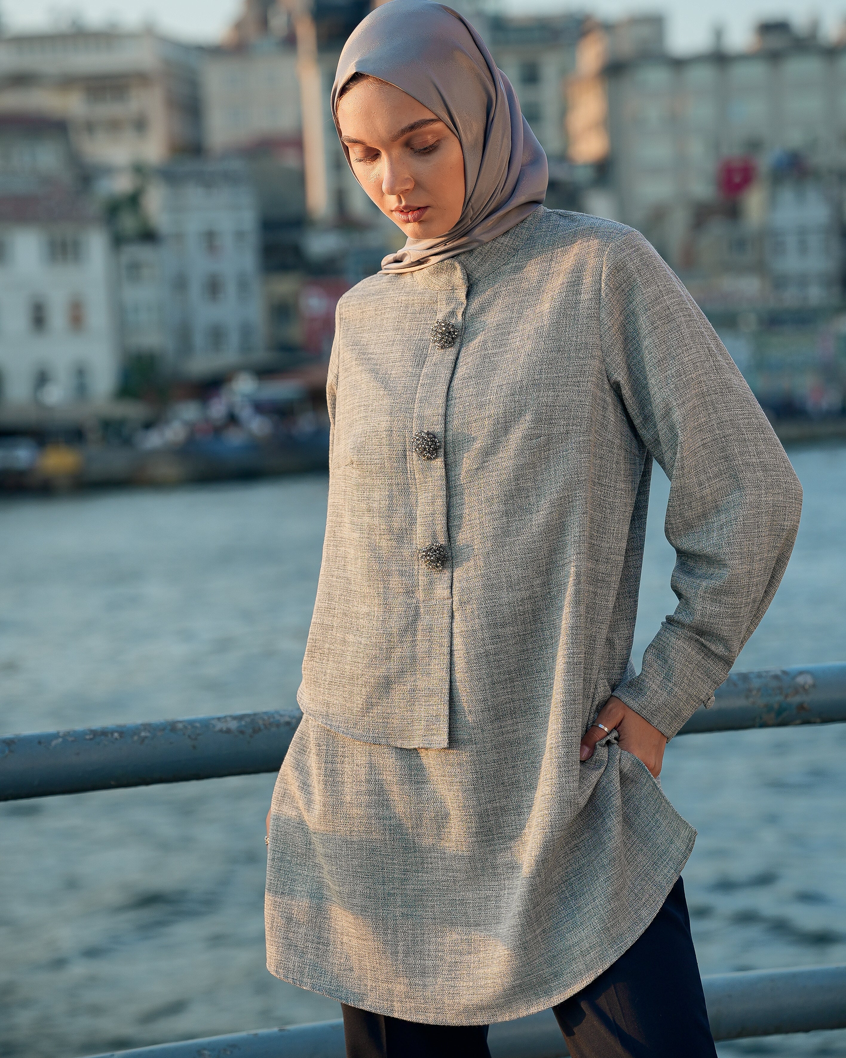 Woman wearing a long, light-colored coat and hijab by a waterfront with buildings in the background.
