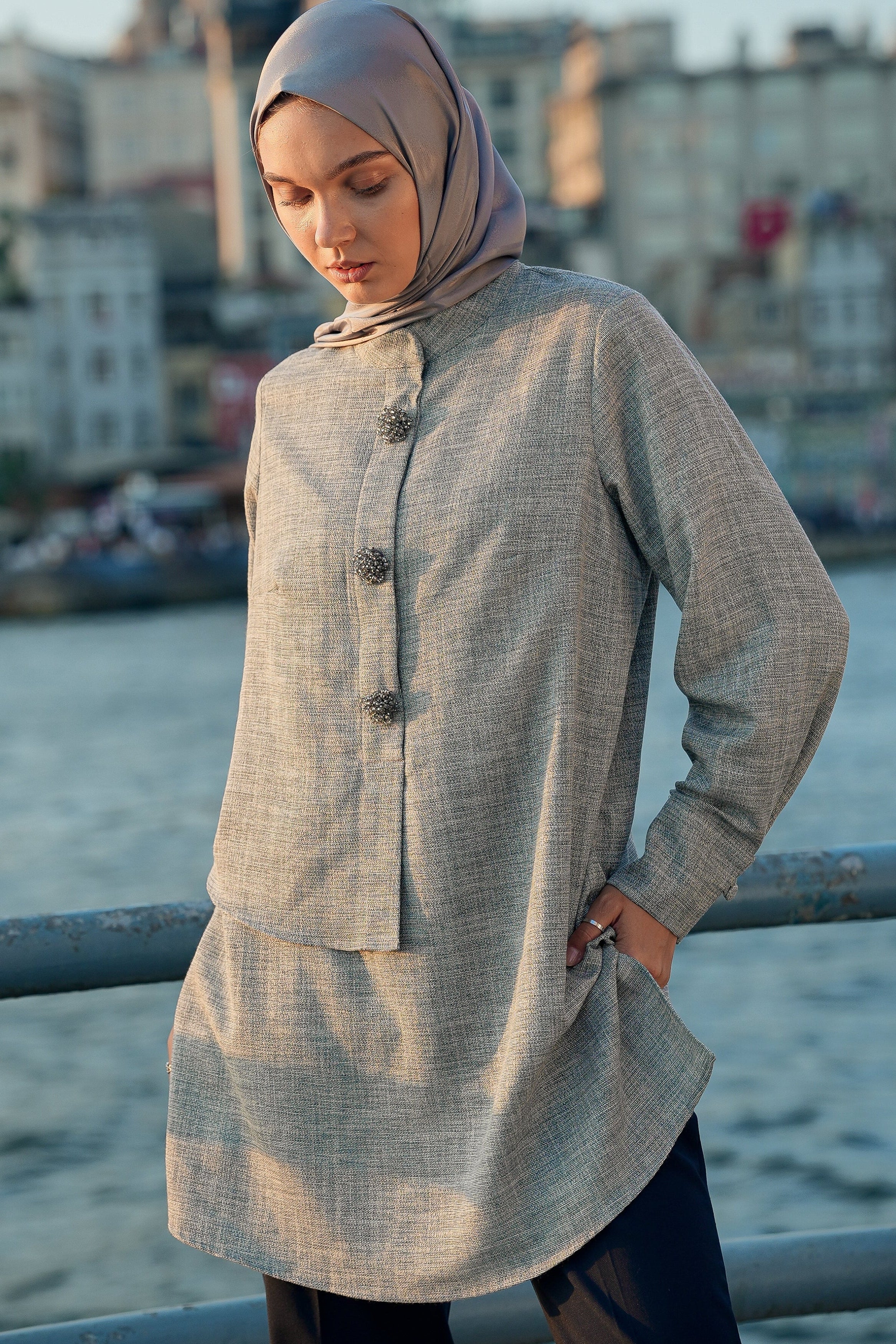 Woman wearing a long, light-colored coat and hijab by a waterfront with buildings in the background.