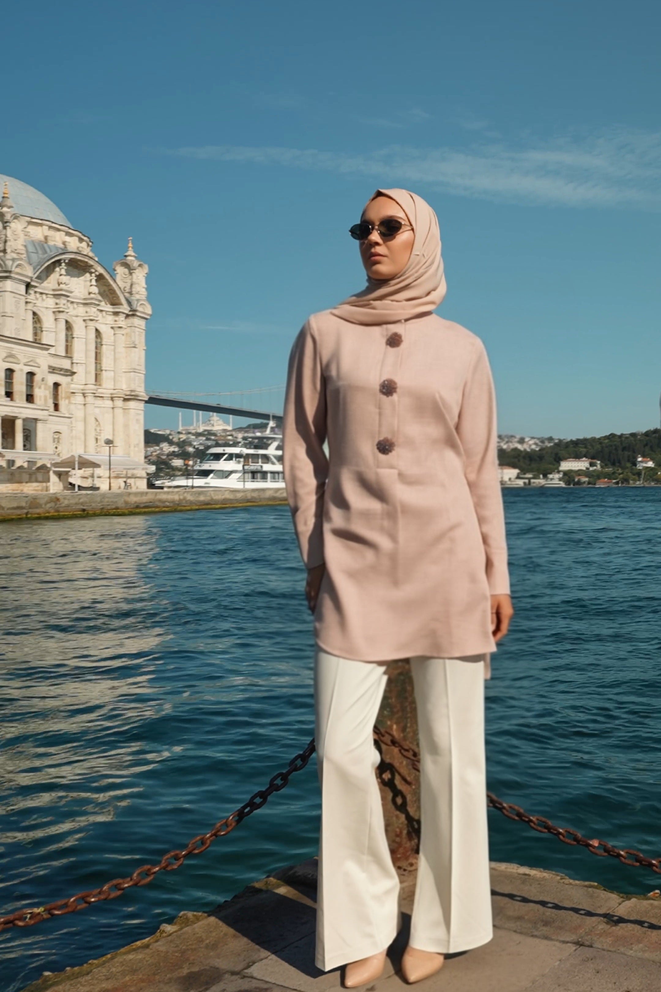 Woman in a light pink hijab and coat standing by a waterfront with a building in the background