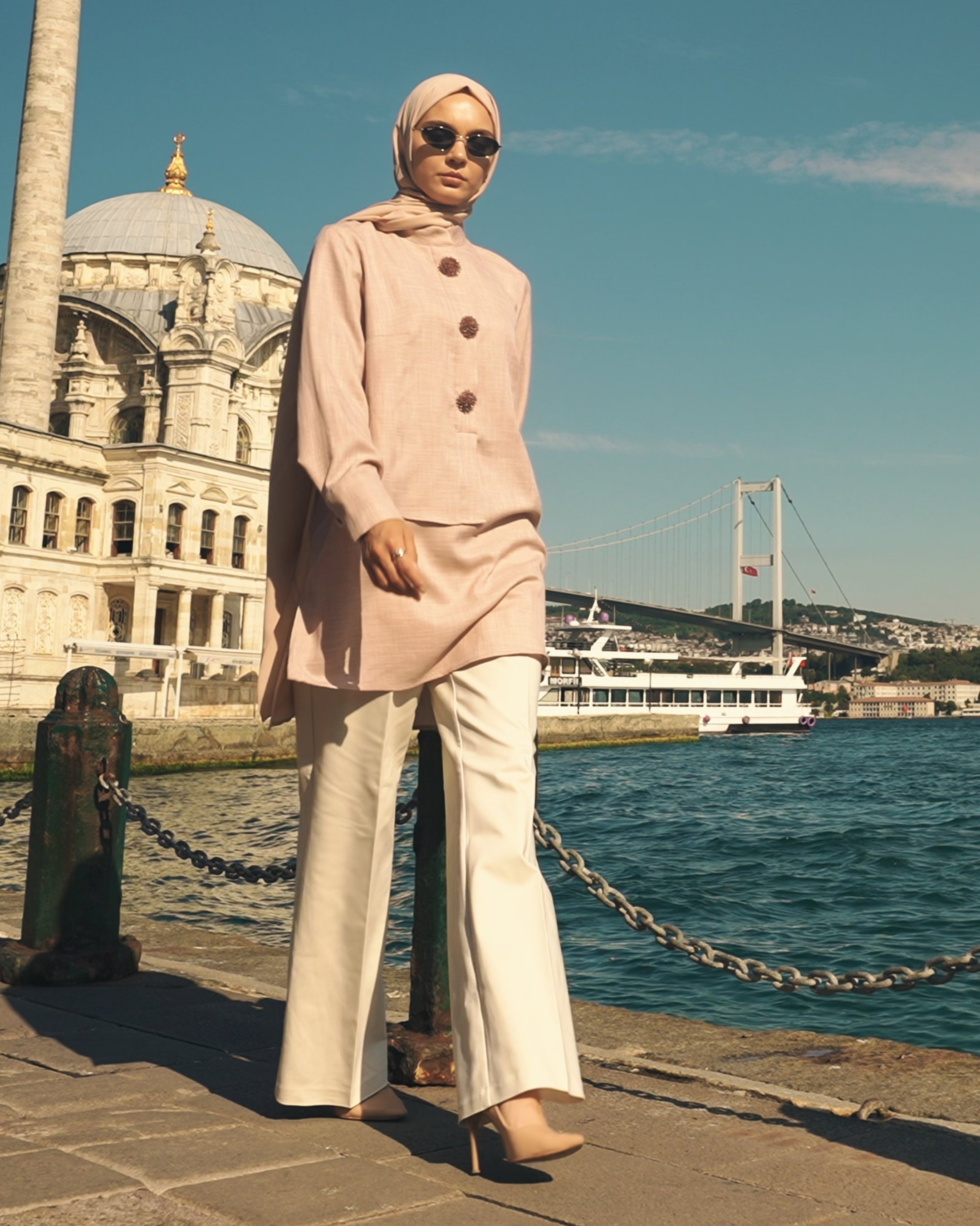Woman in a pink coat and sunglasses standing by a waterfront with a building and bridge in the background.