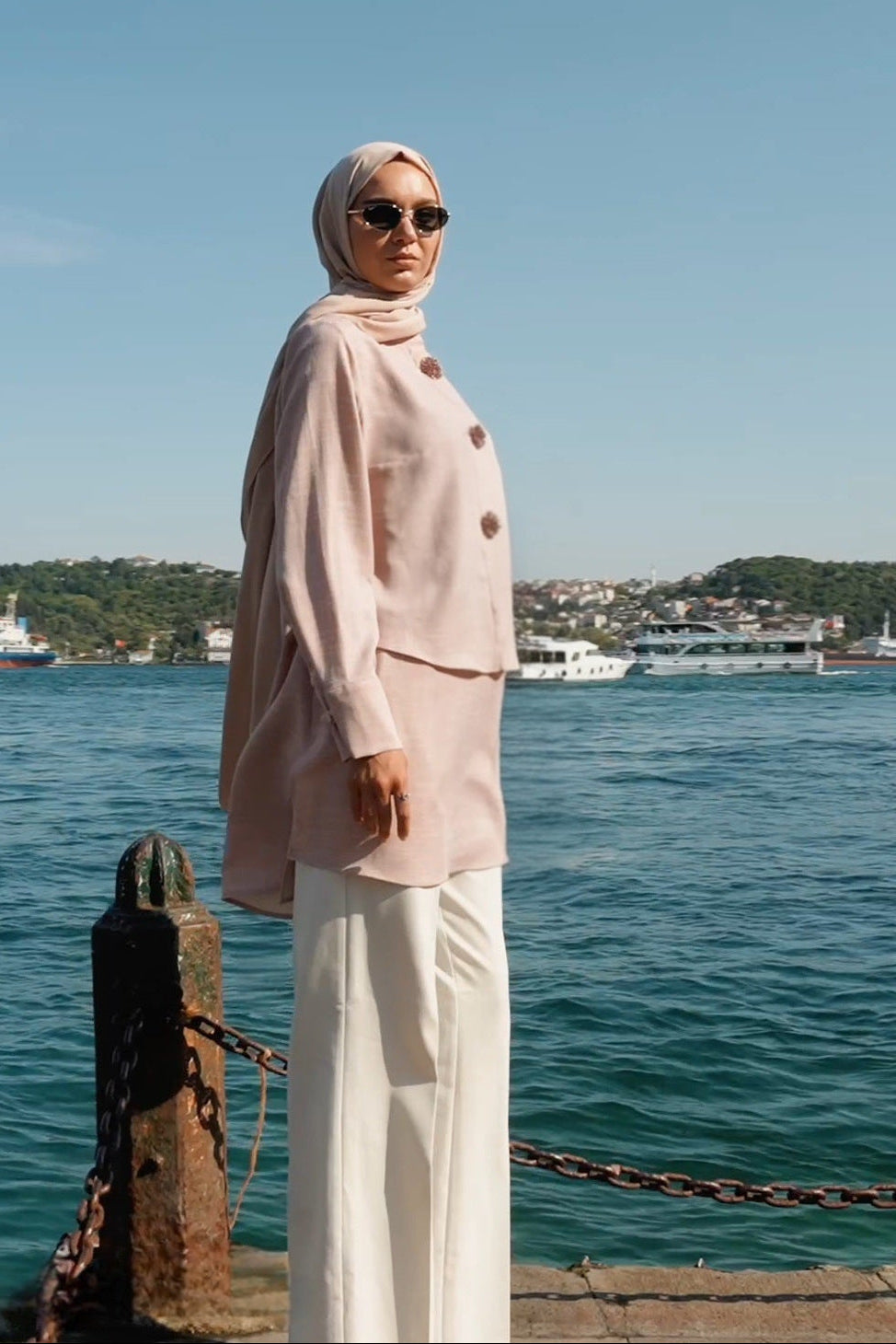 Woman in a light pink outfit standing by a waterfront with a clear blue sky.