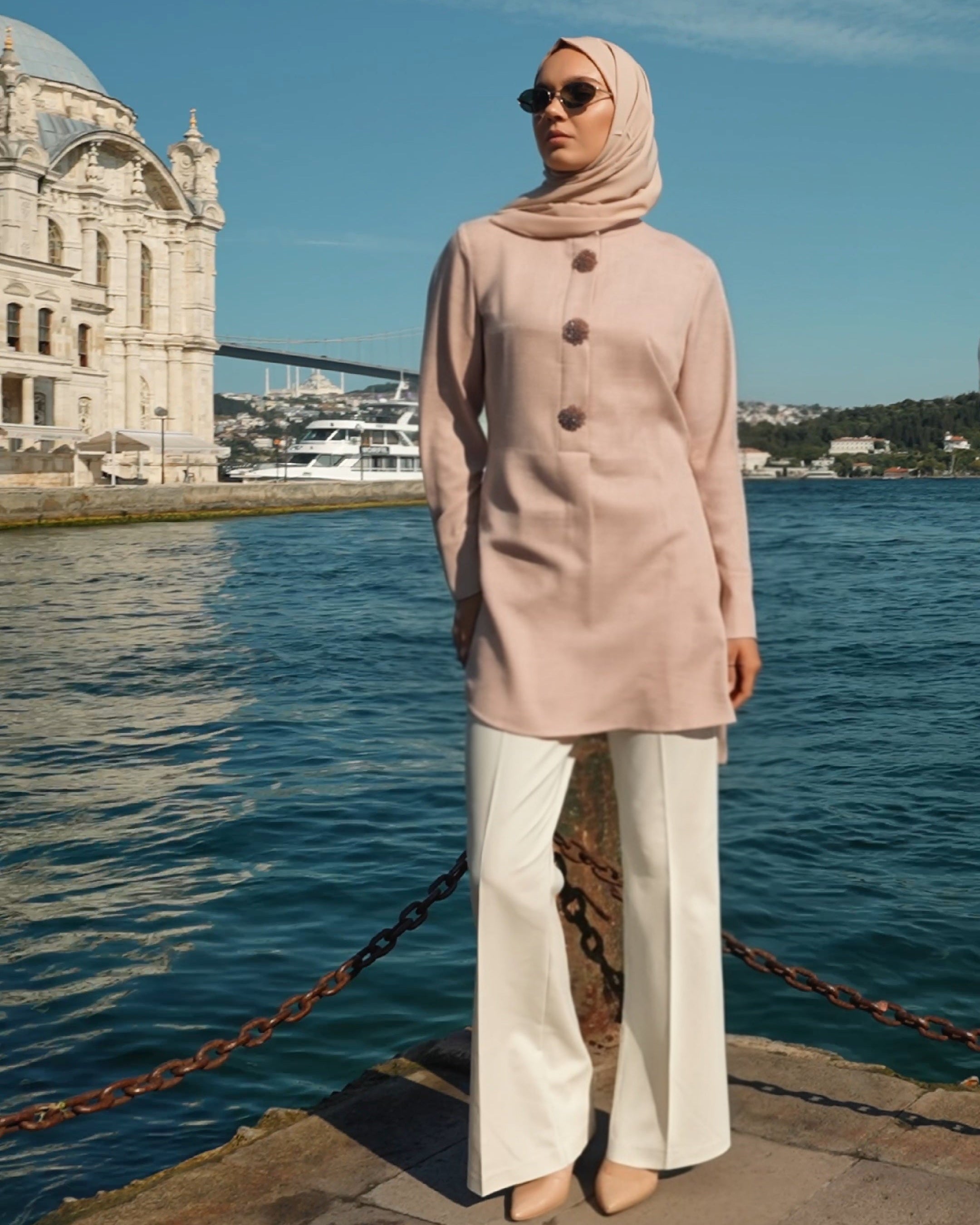 Woman in a light pink hijab and coat standing by a waterfront with a building in the background