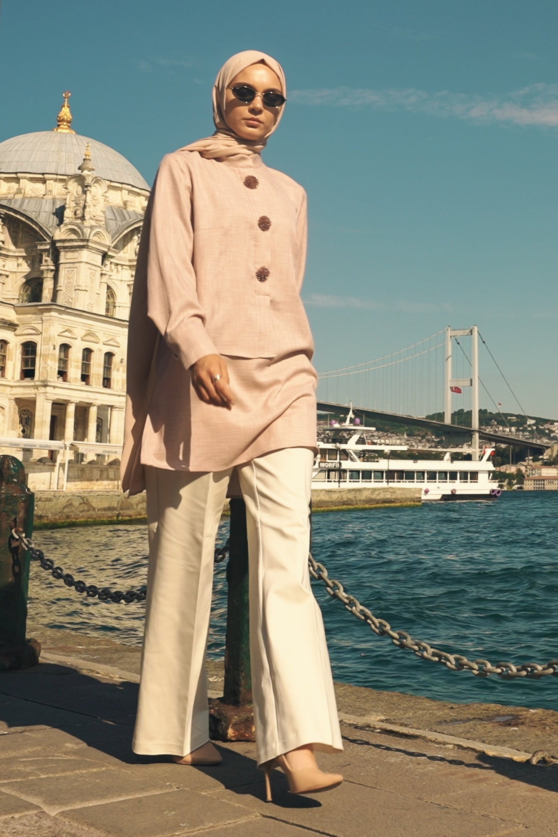 Woman in a pink coat and sunglasses standing by a waterfront with a building and bridge in the background.