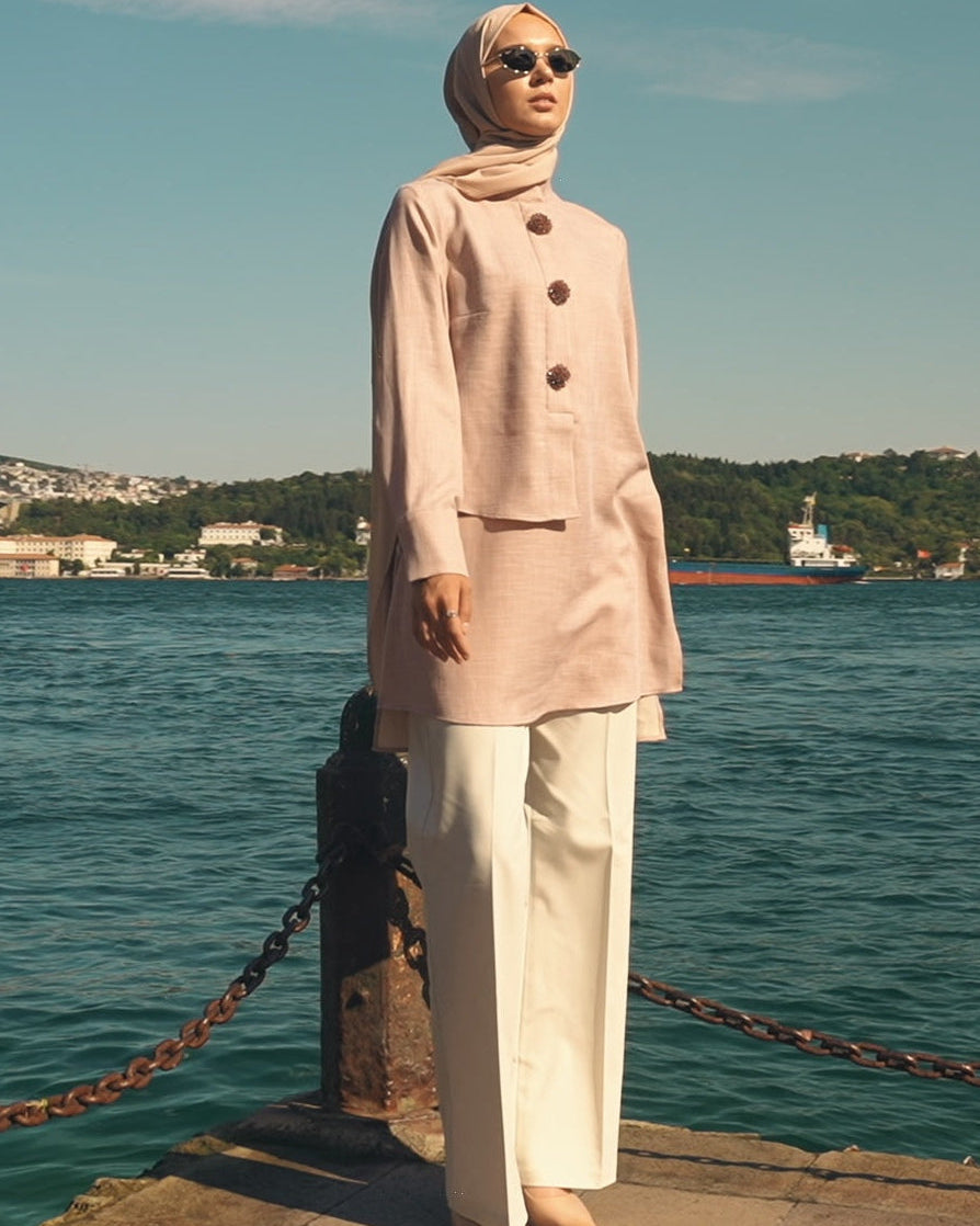 Woman in a light pink coat and hijab standing by a waterfront with a boat in the background.