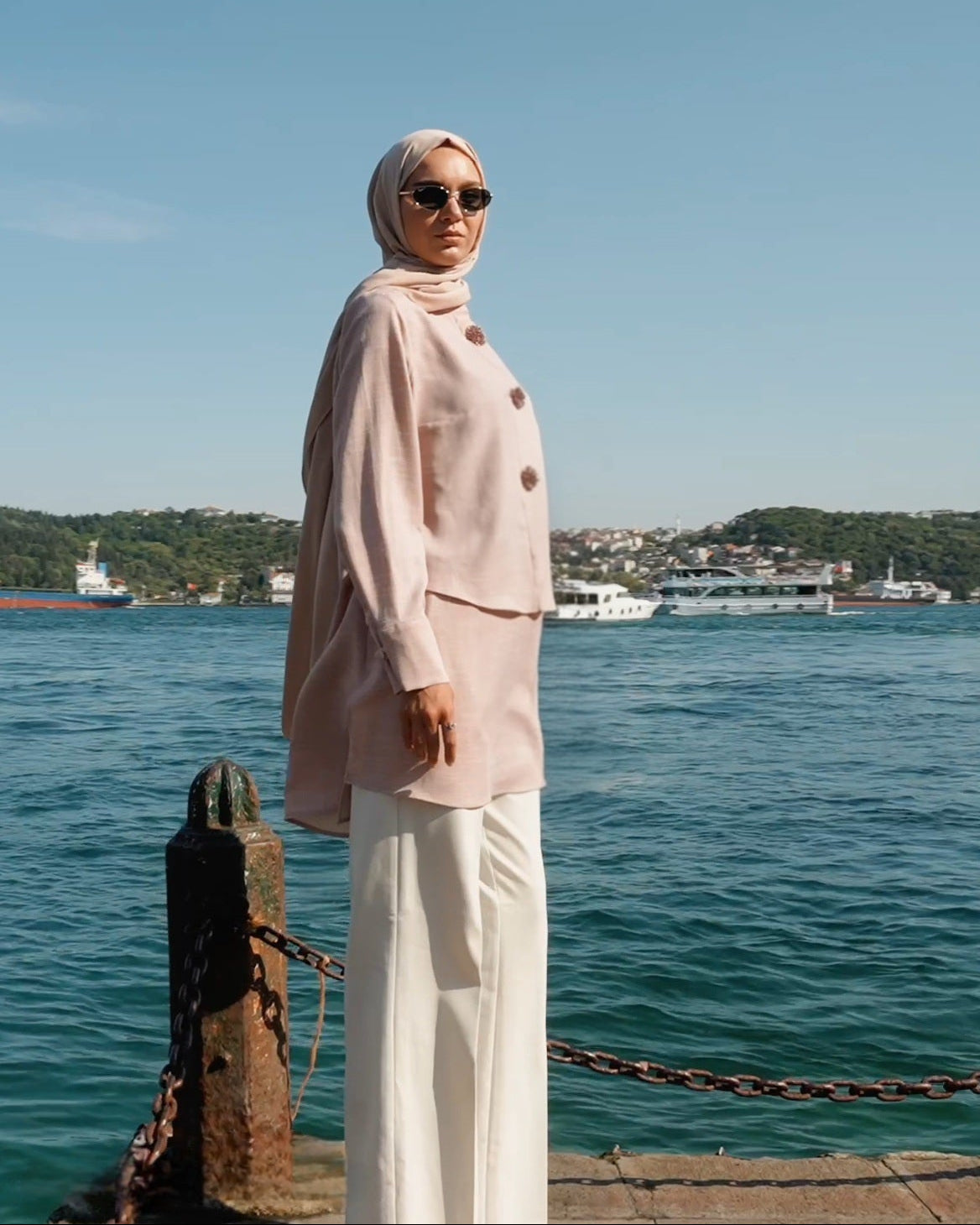Woman in a light pink outfit standing by a waterfront with a clear blue sky.